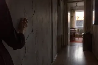 A subtly lit hallway with a partial view of a woman's hand touching a cracked wall, soft yellow light, and moody shadows suggesting an introspective, tense atmosphere.