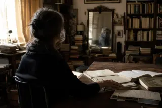 Warm early morning light in a cluttered apartment library, with a woman seen from behind sitting at a table strewn with notebooks and papers, her gaze lost in a mirror's faint reflection.