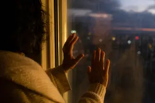 Woman's blurred reflection in a fogged window at dusk, hands pressed on the glass, city lights outside and soft indoor lighting creating an introspective, ambiguous mood.