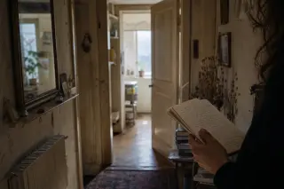 Partial view of a hand holding notes in a softly lit, narrow hallway with cracked walls and morning light streaming through a partially open door.