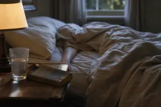 Tangled sheets and a barely visible arm in the soft light of morning on an unmade bed, with a worn book and glass of water beside, evoking introspection and silent psychological tension.