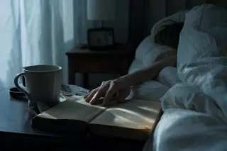Partially visible woman's hand touching an open book on a nightstand in a softly lit, rumpled bedroom at dawn