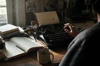 Hesitant hand above a typewriter on an old desk, with open book, erased text, and morning light evoking interrupted thought and introspection