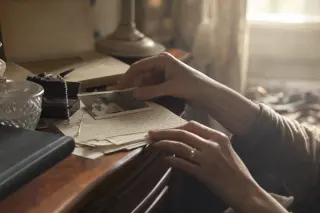 Slender woman's hands touching an old photograph and letters on a cluttered nightstand in soft, shadowed morning light.