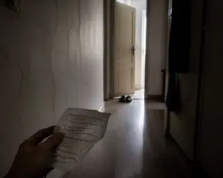 Dimly-lit hallway in an old apartment with pale morning light, a pair of shoes by a half-open door, and the edge of a hand holding crumpled notes in the foreground.