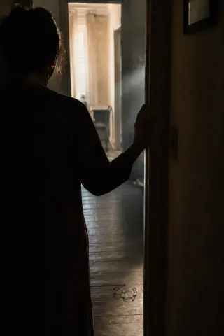 Back view of a woman pausing at a doorway in a dim hallway, with soft morning light and an abandoned pair of glasses on the floor, creating a tense, introspective atmosphere.
