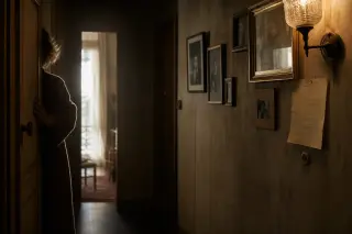 Hand of a middle-aged woman on an open door in a softly lit, narrow hallway with faded family photos and a handwritten note on the wall, captured in early morning light with a quiet, introspective mood.