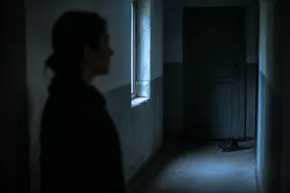 Middle-aged woman seen from behind in a dim, narrow corridor, pausing by a fogged window with soft morning light and faint silhouettes outside, conveying a sense of introspective transition.