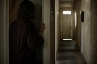 A pair of mature woman's hands hesitates over an open book with a faded note, in a softly lit, cluttered library at dusk.