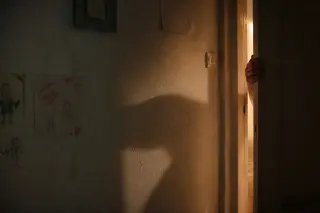 A woman's hand on a doorframe in a dim hallway, warm light casting shadows and faded childhood drawings visible on a cracked wall.