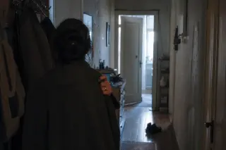 Woman's back in a dimly lit, old hallway at dawn, hand pausing on a coat, soft natural light casting long shadows, mood of subdued anticipation.