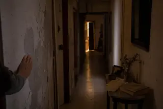 Dimly lit corridor at dusk with a woman's hand on a cracked wall, open door in distance, handwritten notes and book on table, warm filtered light, introspective atmosphere